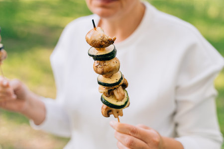 Mature woman 50 years old eating grilled bbq vegetables from wooden skewers outdoors in summer forestの写真素材