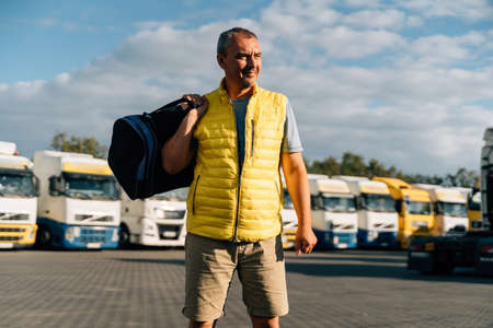 Portrait of caucasian mature man with bag on some-truck vehicles parking background. Truck driver workerの写真素材