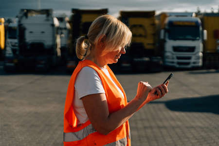 Happy middle-aged woman having a phone call in front of yellow semi-truck vehicleの写真素材
