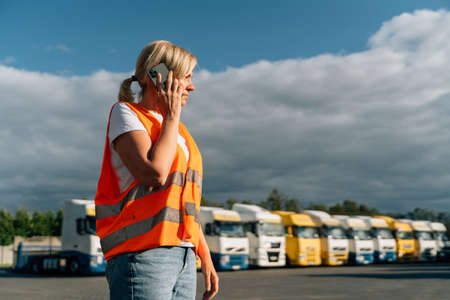 Happy middle-aged woman having a phone call in front of yellow semi-truck vehicleの写真素材