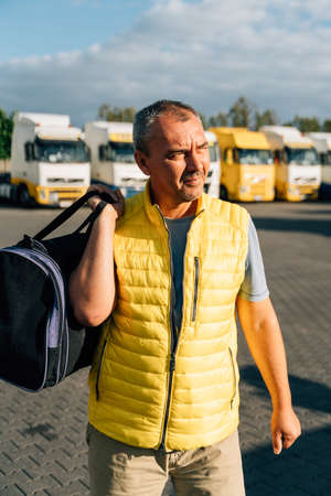 Portrait of caucasian mature man with bag on some-truck vehicles parking background. Truck driver workerの写真素材
