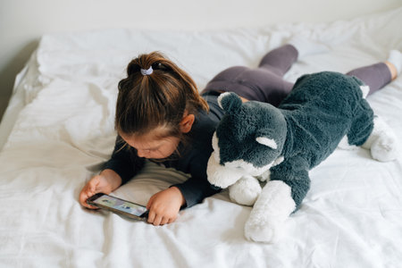 preschool age girl watching cartoons on smartphone on bed. Plush toy dogの写真素材