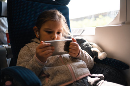 Child girl traveling by train and watching cartoons on phone with wireless headphonesの写真素材