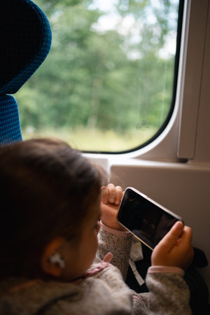 Child girl traveling by train and watching cartoons on phone with wireless headphonesの写真素材