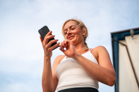 Beautiful blonde caucasian woman using phone while doing sports outdoors. Healthy fitness lifestyle, summer workout in city. Using technology for tracking activityの写真素材