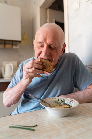 Elderly senior man eating soup with bread and green onion at home in the kitchen. Happy retirement, simple whole food.の写真素材