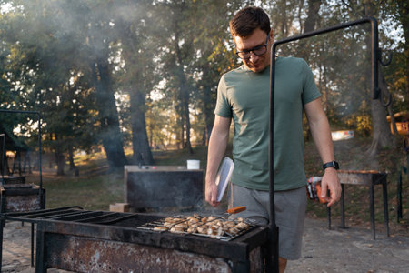 Middle age caucasian man blowing air into the bbq coals while cooking grilled champignons. Outdoors BBQ party, simple tasty foood on fireの写真素材