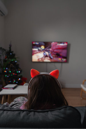 Back view of a child girl sitting on the couch and playing video game or watching cartoons on tv. Wireless funny pink headphones with earsの写真素材