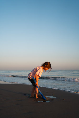 A young child gazes into the sea while holding a blue shovel on the beach, with the warm glow of sunset lighting the serene sceneの写真素材