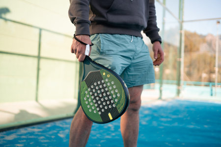 Close-up of a mans hands gripping a padel tennis racket, ready to play, with the courts fence and blue surface in the backgroundの写真素材