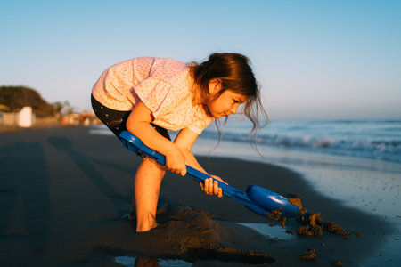 Intently focused, a young child digs in the sand with a blue shovel on the beach, with the evening light casting a soft glow on the activityの写真素材