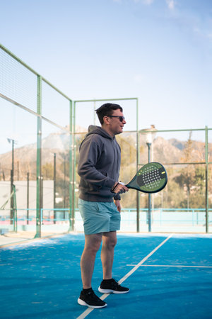 A man in a hoodie and shorts smiles while holding a padel tennis racket on a sunny outdoor court with mountains in the distanceの写真素材