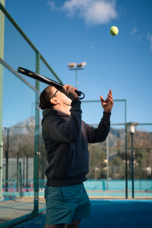 A man in sunglasses serves a tennis ball, enjoying a paddle tennis match on a sunny outdoor court...