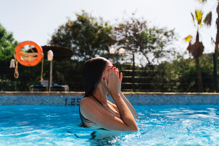 A young woman playfully splashes water in a pool, covering her face with her hands, having fun under...
