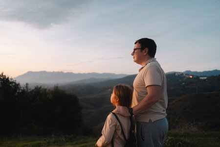 A father embraces his daughter as they enjoy the scenic view of hills and mountains under an evening sky.の写真素材