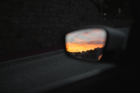A cars side mirror capturing the vibrant colors of a sunset, with a stone wall and road in the backgroundの写真素材