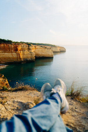 A personal perspective of a person relaxing on the cliffs of the Algarve, Portugal. Golden limestone cliffs meet tranquil blue waters under a bright, sunny sky.の写真素材