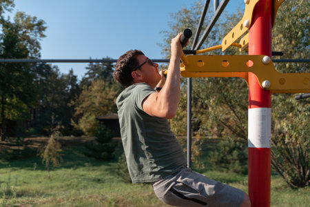 A man focuses on pull-ups during a workout on outdoor gym equipment, surrounded by nature on a sunny dayの写真素材