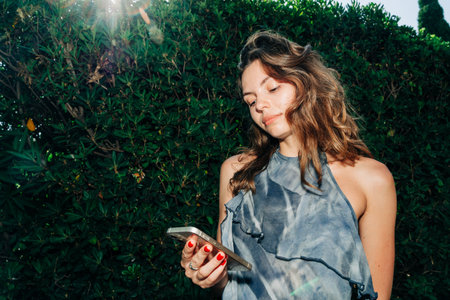 A smiling woman holds up her smartphone to capture a selfie, standing against lush green bushes,...
