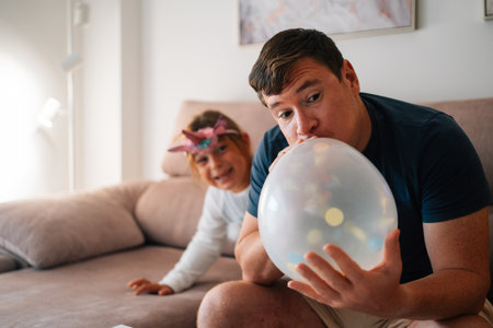 Caucasian male adult inflating balloon with hispanic child in playful indoor settingの写真素材