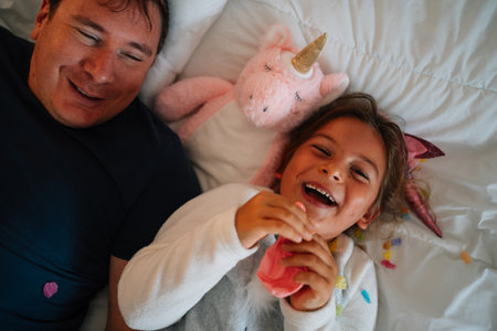 Joyful caucasian adult male and child laughing with unicorn toy on bed