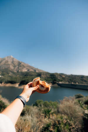 Hand holding sandwich with mountain and lake view under clear blue skyの写真素材