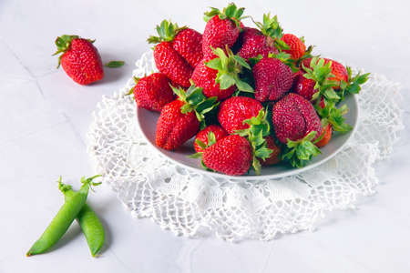 Fresh ripe juicy red strawberries on white plate on white doily and pods of green peas on white marble table.の写真素材