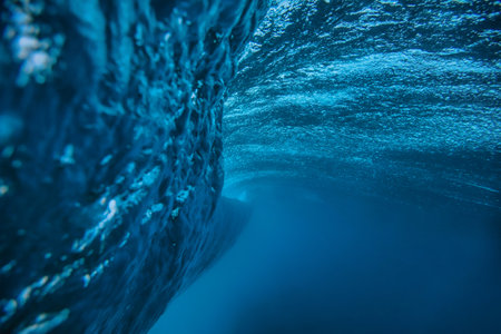 Underwater view breaking ocean wave tunnel deep blue barrel curling above, light filtering through translucentの写真素材