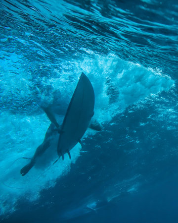 Surfer duck dives under glassy wave, underwater view shows surfboard nose cutting through blue water, sunlightの写真素材