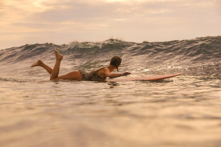 Young Woman Paddling On Surfboard At Sunset, Lying Prone And Stroking Toward Horizon, Warm Golden...
