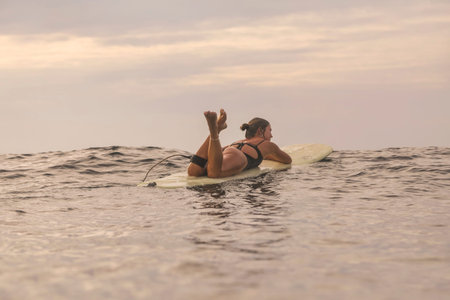 Young Woman Resting On Surfboard During Sunset Lull, Legs Playfully Raised, Soft Waves And Warm Sky,...