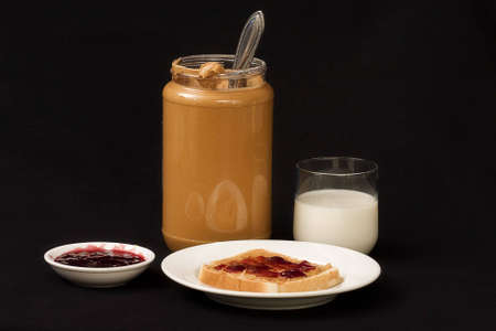 Large jar of peanut butter and dish of jelly along with plate with sandwich and milk beside it.の写真素材