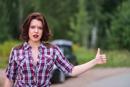 Beautiful young woman hitchhiking gesture at countryside, broken car at background.の写真素材