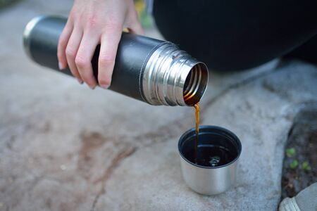 Young woman, sitting on a big stone preparing hot tea from a thermos into a cap.