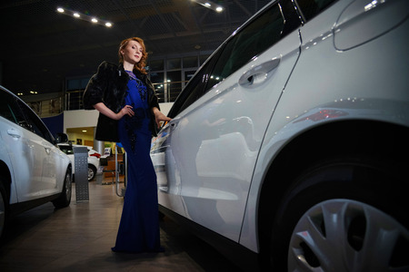 Elegant woman with red hair in blue dress and fur coat standing near new modern car.の写真素材