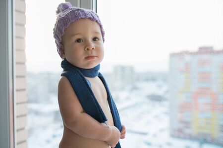 Happy little baby boy standing near the window weared in winter hat and scarf, city at backgroundの写真素材