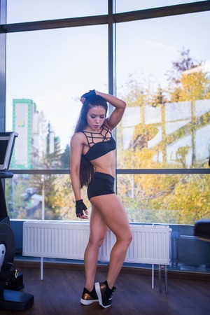 Young sexy woman with beautiful body and long dark hairs posing near panoramic window in fitness club. City, autumn yellow trees at background.の写真素材