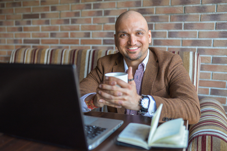 Happy Indian man sitting at cafe with notebook and laptop with cup of coffee, smiling looking at camera.の写真素材