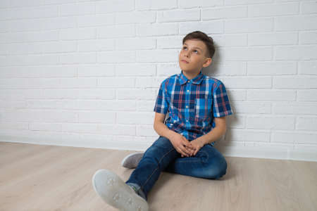 Young boy thinkin sitting near white white brick wall looking up, copy space available.の写真素材