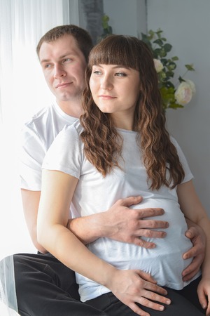 Young pregnant woman and her husband are hugging, touching the belly near window and looking ousideの写真素材
