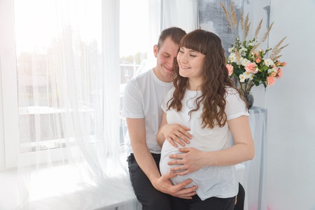 Happy young pregnant woman and her husband embracing, touching the belly near window and smilingの写真素材