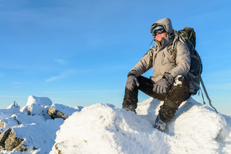 Hiker with backpack sitting and having rest on the top of a snow-covered rock over the winter mountainsの写真素材