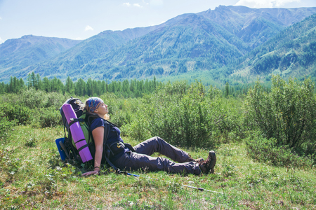 Woman hiker with backpack sitting on green grass and feel relax with closed eyes at background of high mountainsの写真素材