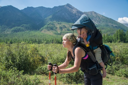 Tired Woman Hiker trekking in mountains with child in backpack, stopped for rest. Mother with baby boy travelling in summer sunny day.の写真素材
