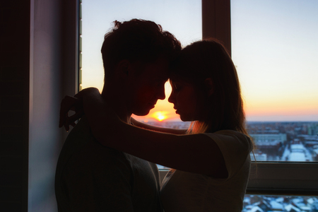 Couple in love, profile silhouettes close to each other, beautiful sunset in window at background.の写真素材