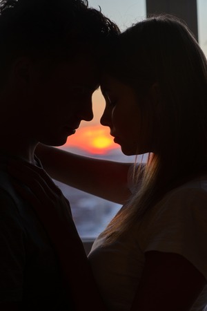 Couple in love, profile silhouettes close to each other, beautiful sunset in window at background.