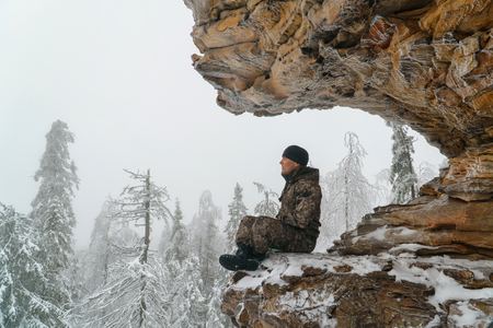 Man hiker sitting on scenic rock in meditating relax pose, winter mountains and fir forest covered with snow at background.の写真素材