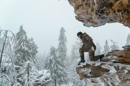 Man hiker looking down from high altitude standing on scenic rock, winter mountains and fir forest covered with snow at background.の写真素材