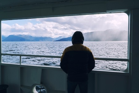 Man in warm jacket with hood on ferry looking at sea and mountains of Norway.の写真素材