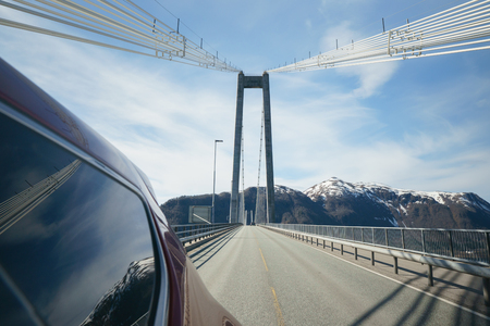 View back from side of car, driving on modern bridge at sunny day with beautiful blue sky and mountains at background. Travelling in Norway.の写真素材